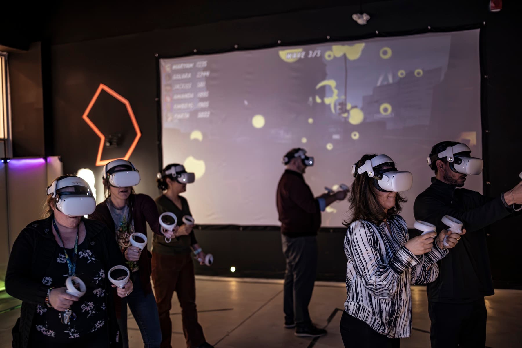 Six coworkers in VR headsets playing a team game with live scores on a projection screen